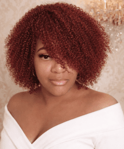 Woman wearing a burgundy Jerry curl human hair bob wig with defined deep wine red curls, styled in an elegant off-shoulder white top against a luxury cream background.